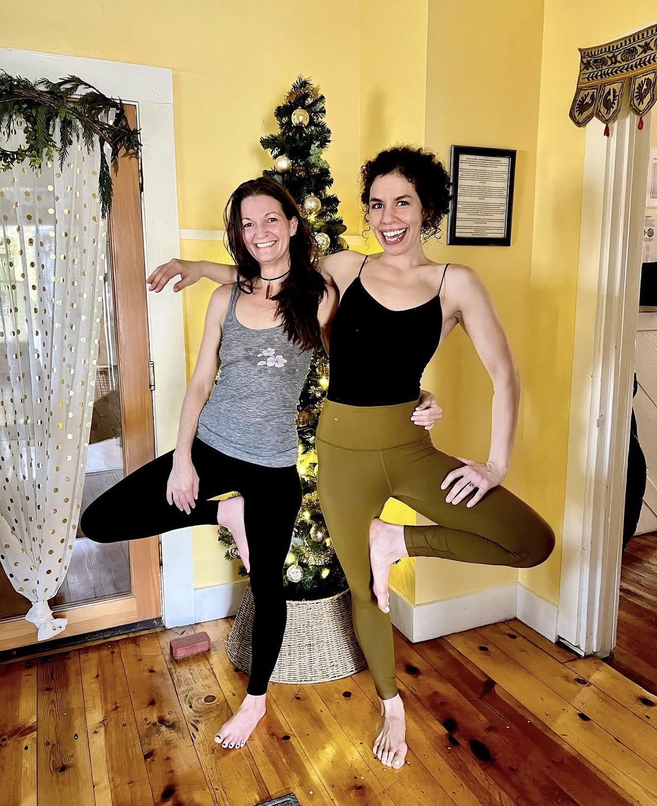 Two teachers in tree pose beside the holiday tree